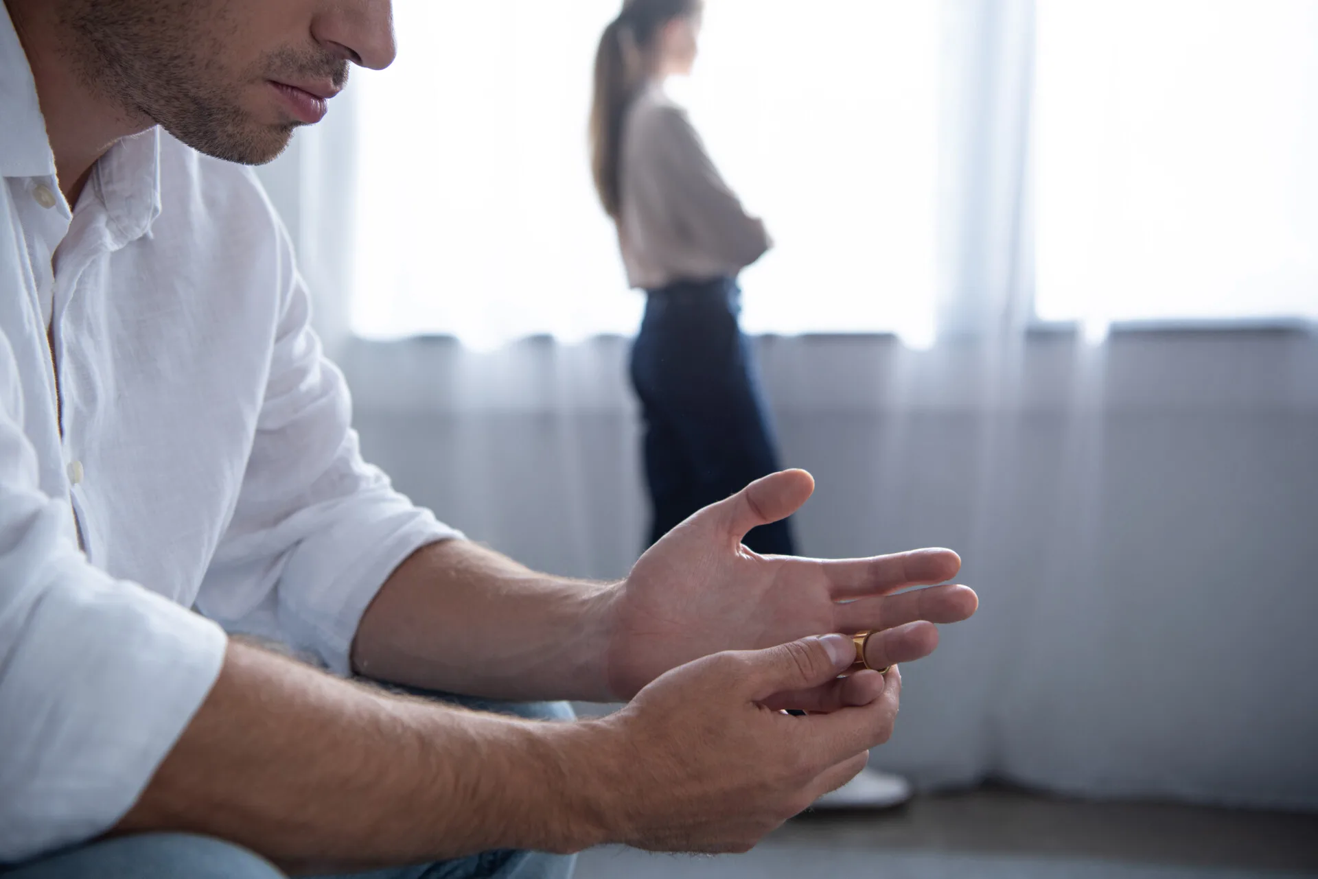 Kvinde står ved vinduet, mens mand sidder og piller ved sin ring
